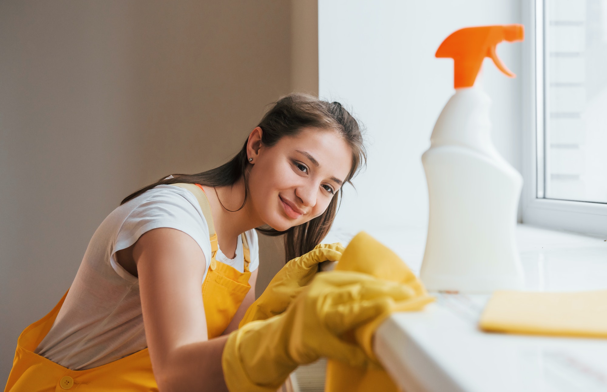Housewife in yellow uniform works with window and surface cleaner indoors